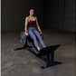 Woman in burgundy tank top and blue leggings using black leg press machine in industrial gym setting