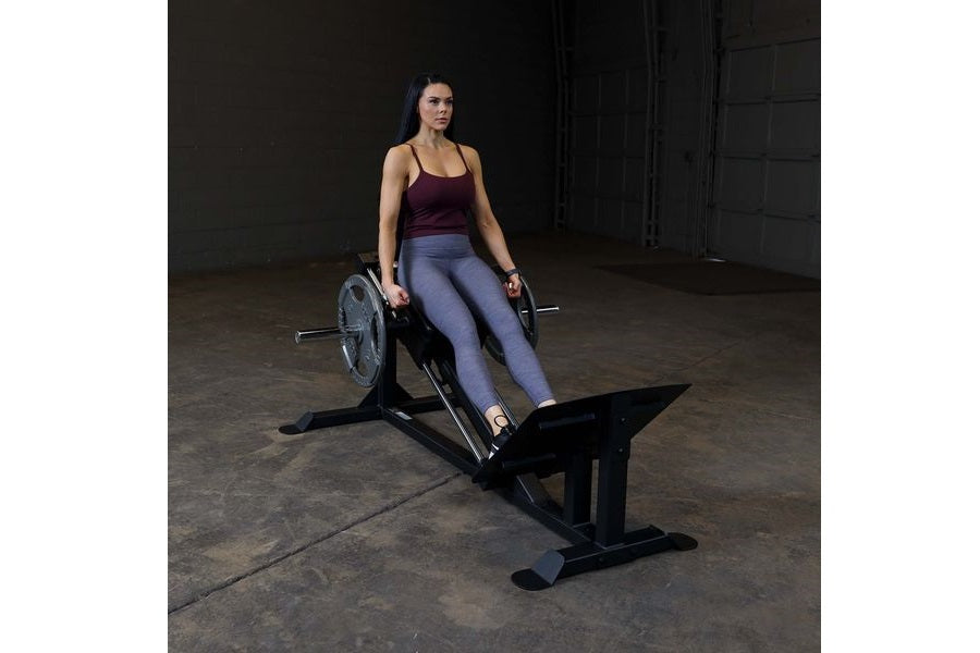 Woman in burgundy tank top and blue leggings using black leg press machine in industrial gym setting