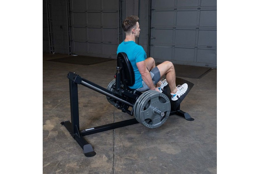 Man wearing blue t-shirt using black Body-Solid leg press machine with weighted barbell in garage gym setting