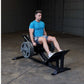 Man in blue t-shirt and gray shorts using black Body-Solid leg press machine with weight plates in industrial gym setting