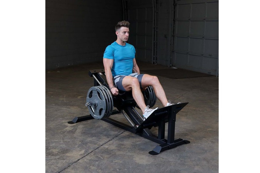 Man in blue t-shirt and gray shorts using black Body-Solid leg press machine with weight plates in industrial gym setting