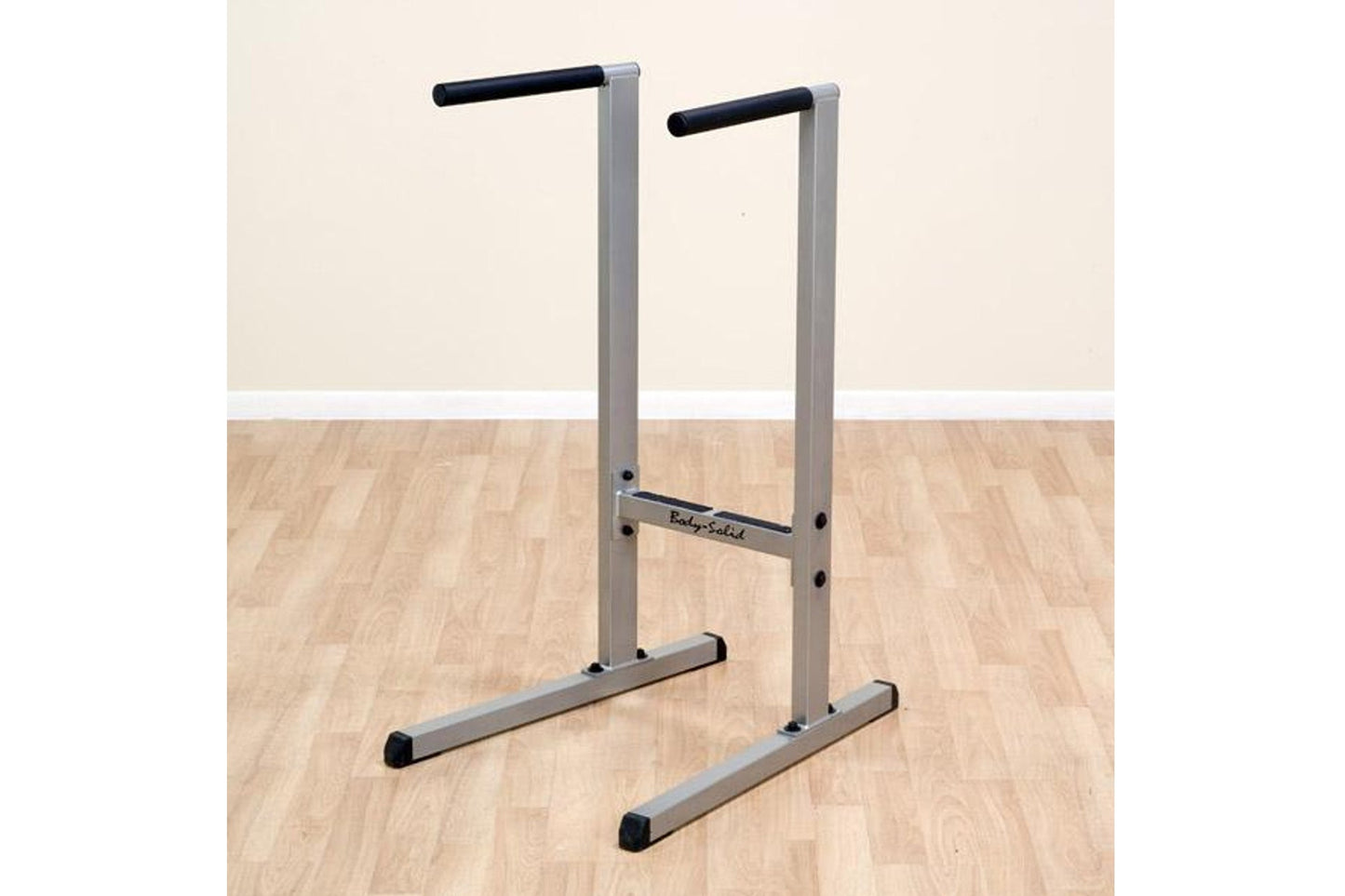 Body-Solid dip station with gray aluminum frame, black handles, and stabilizing feet on wooden floor