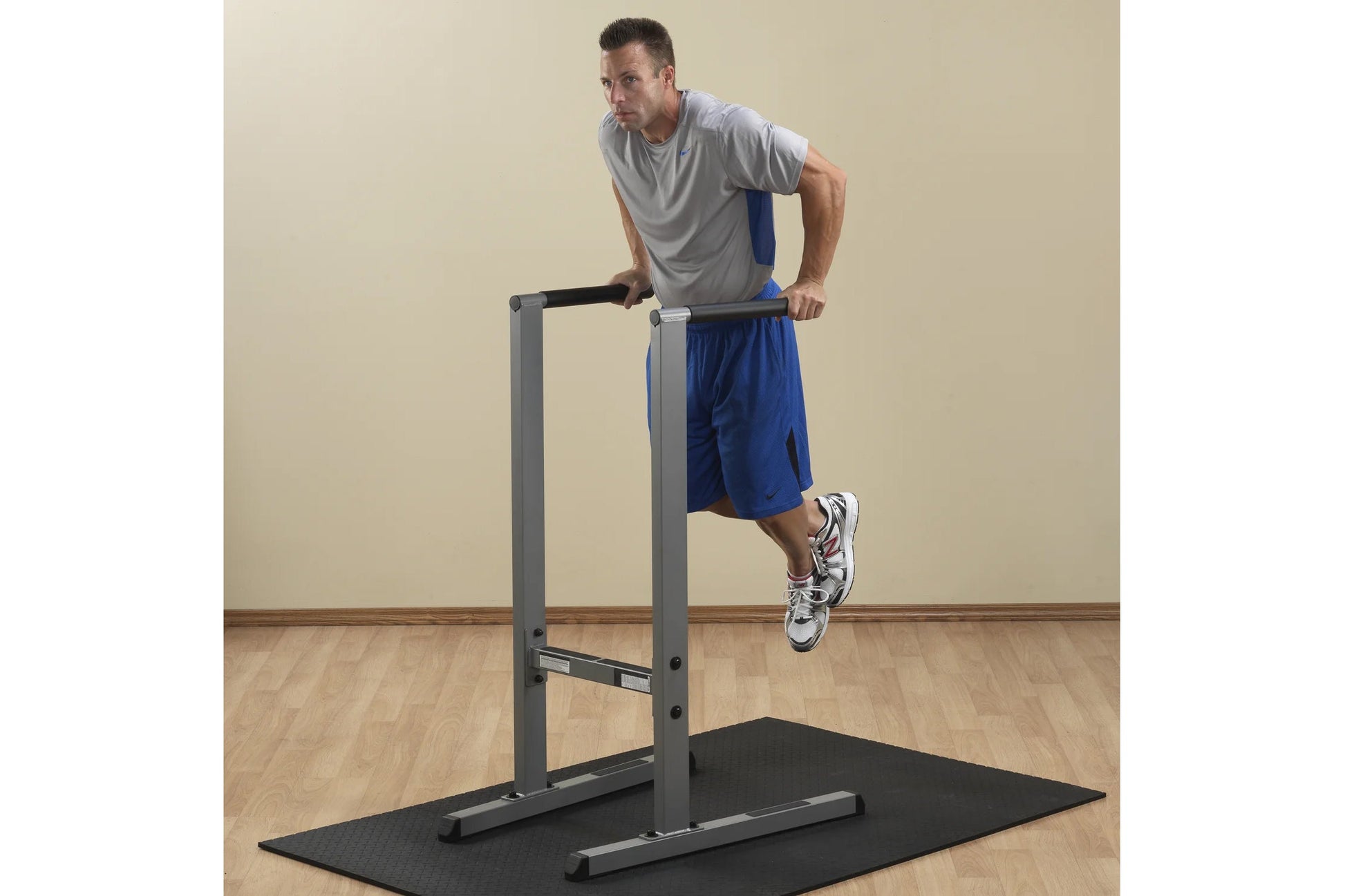 Man using gray dip station with black base mat, wearing gray t-shirt and blue shorts on wooden floor