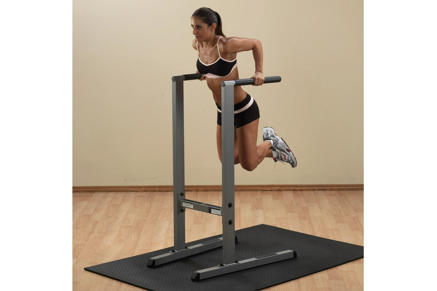 Woman using gray and black dip station with pull-up bar on exercise mat, wearing black sports bra and shorts with white trim