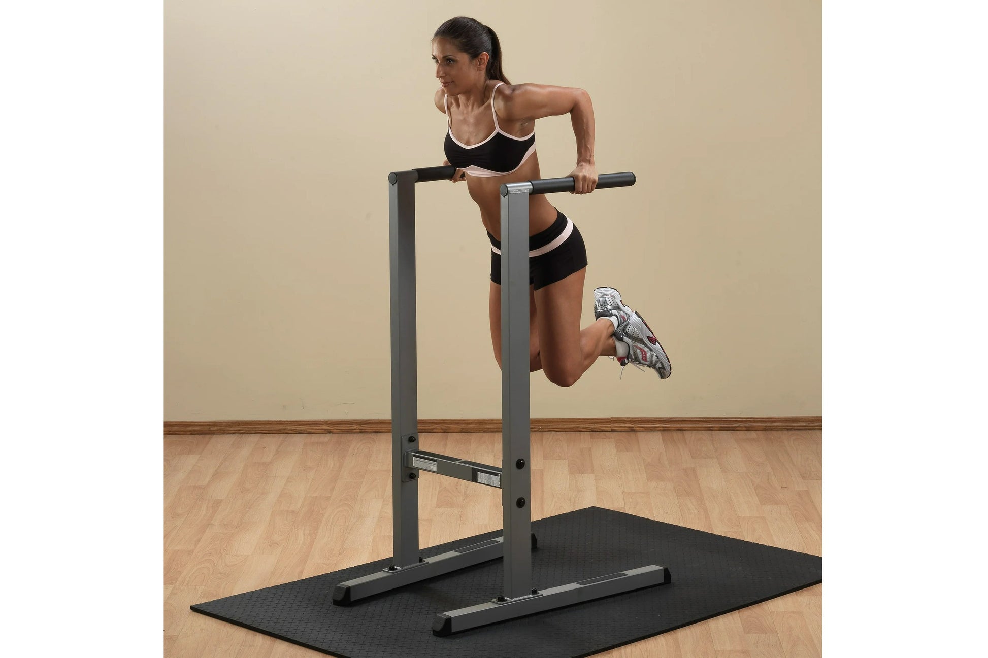 Woman using gray and black dip station with pull-up bar on exercise mat, wearing black sports bra and shorts with white trim