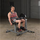 Man in red striped tank top performing leg extension on gray and black weight machine with dumbbells in gym setting