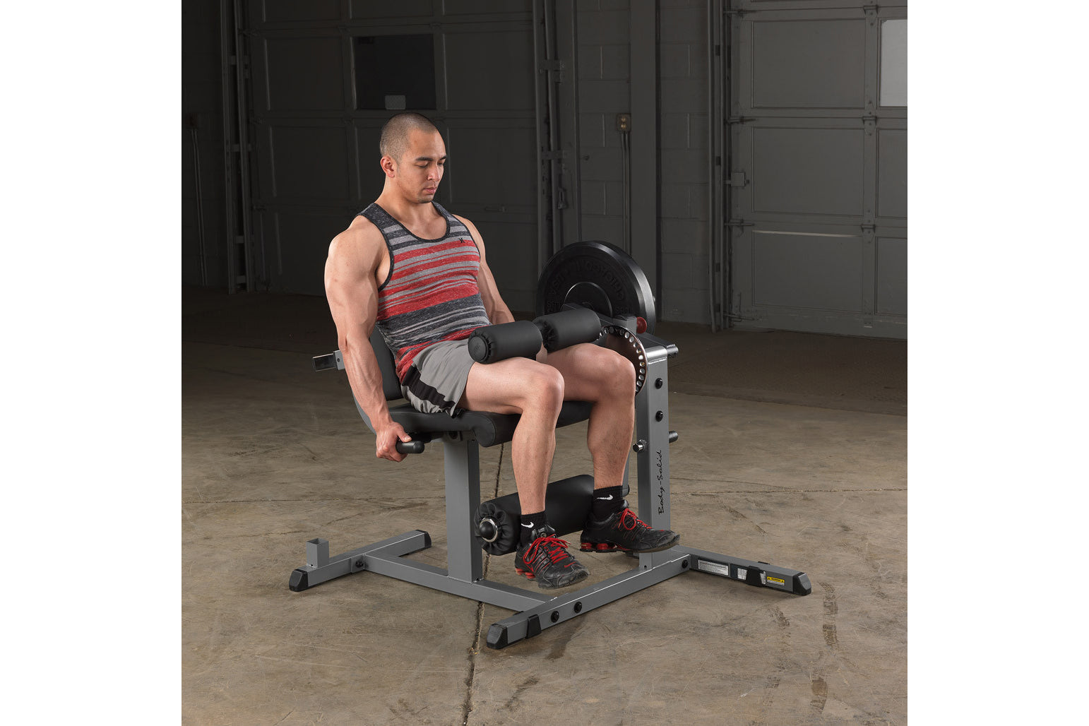 Man in red striped tank top performing leg extension on gray and black weight machine with dumbbells in gym setting