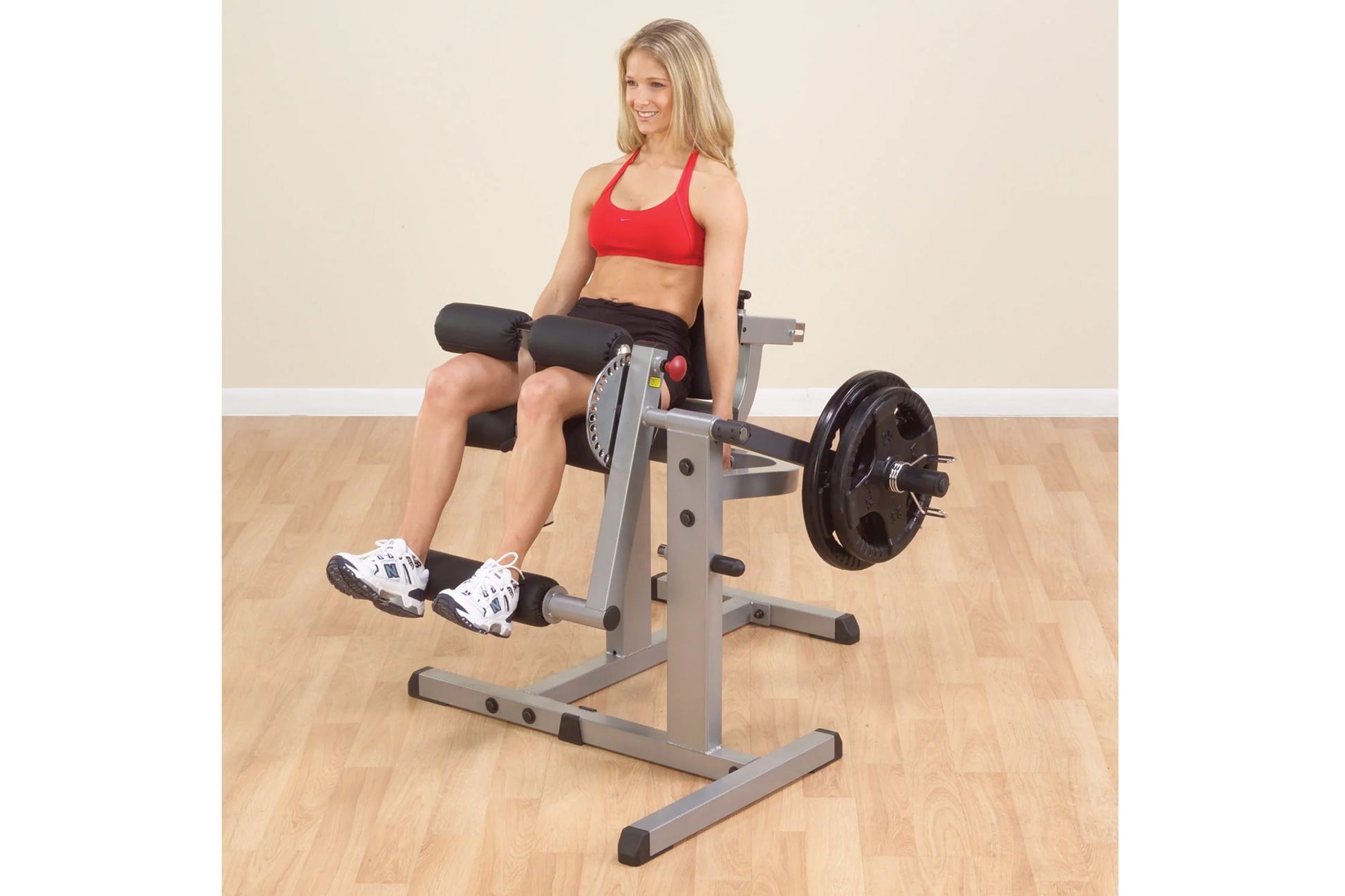 Woman in red sports bra and black shorts using gray and black leg extension machine with weight plate in gym setting