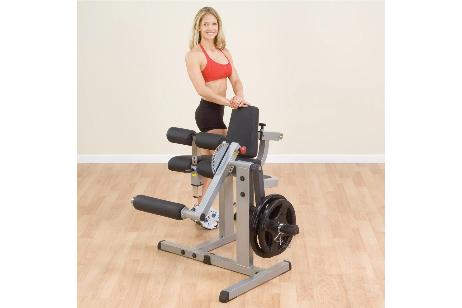 Woman in red sports bra and black shorts seated on Body-Solid leg extension and curl machine with weight plate, shown in gym 