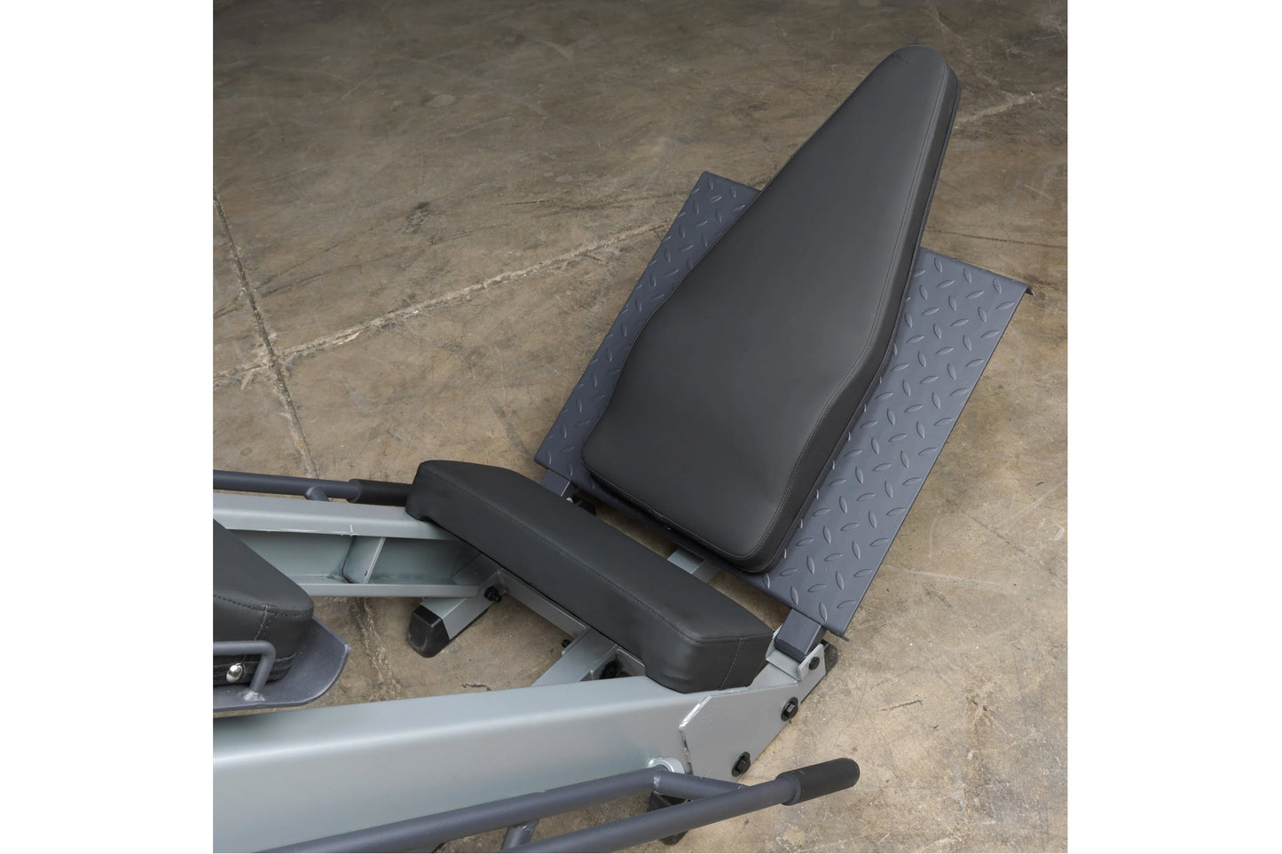 Black padded seat with blue textured footplate on silver Body-Solid leg press machine, viewed from above on concrete floor