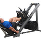 Man in blue shirt using gray and black leg press machine with loaded weight plate, demonstrated on white background