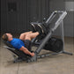Gray and black leg press machine with loaded weight plates, man in blue shirt demonstrating exercise in gym setting