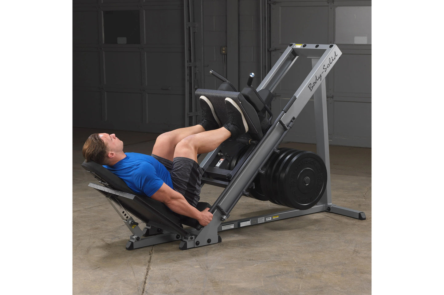 Gray and black leg press machine with loaded weight plates, man in blue shirt demonstrating exercise in gym setting