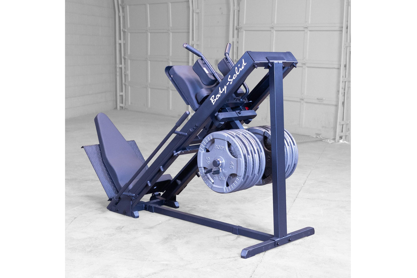 Body-Solid leg press and hack squat machine in navy blue with padded seat, foot platform, and weight plate storage on concret