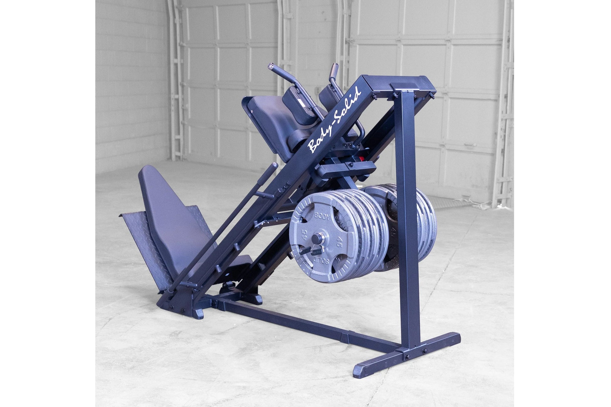 Body-Solid leg press and hack squat machine in navy blue with padded seat, foot platform, and weight plate storage on concret