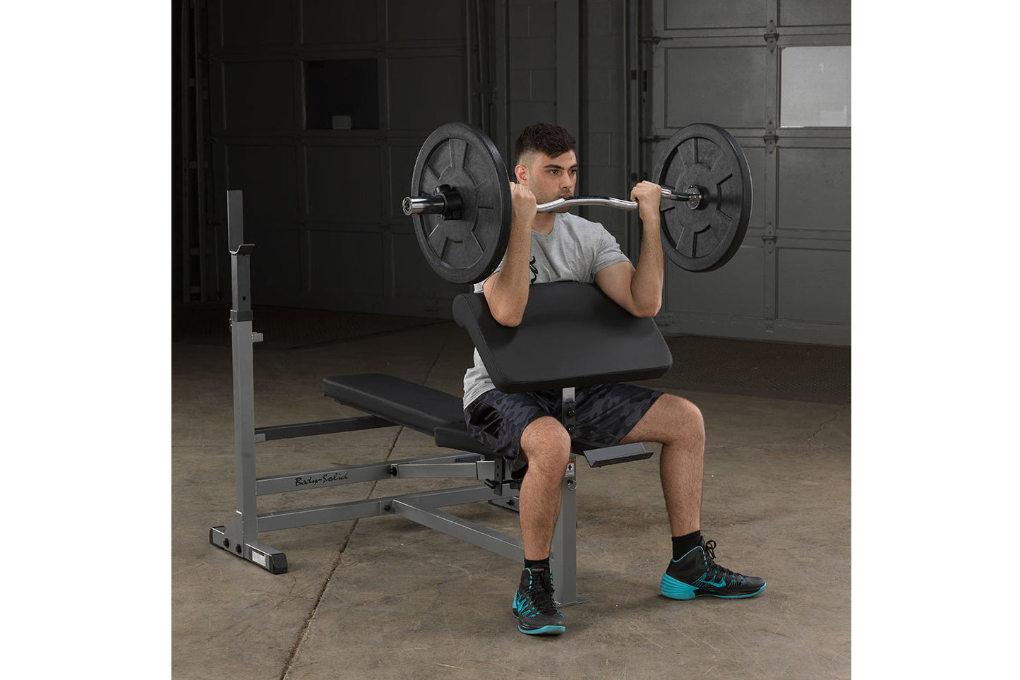Man performing barbell shoulder press on black Body-Solid weight bench with loaded barbell in industrial gym setting