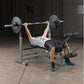 Person performing barbell bench press on gray and black weight bench with leg extension attachment in industrial gym setting