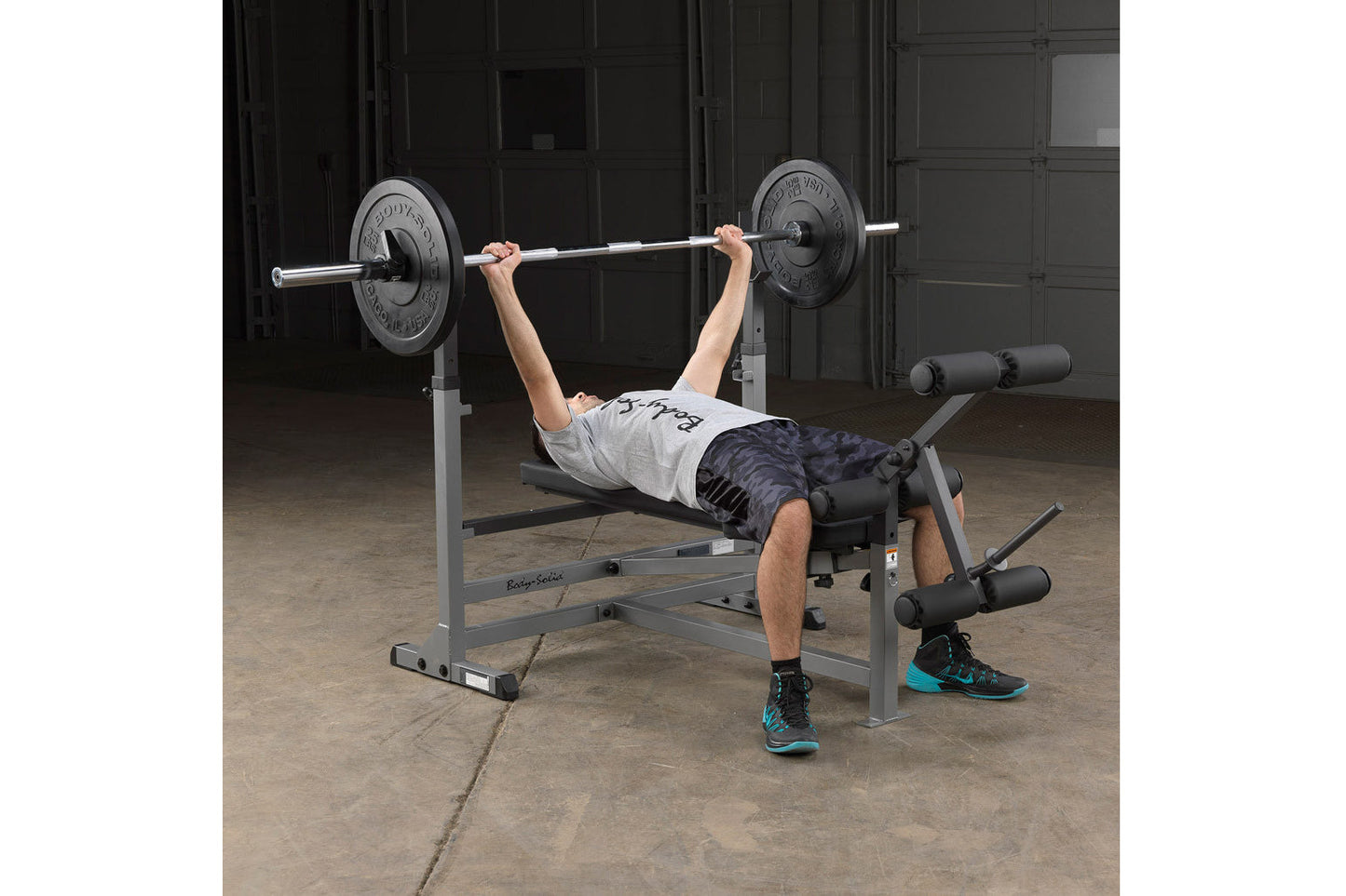 Person performing barbell bench press on gray and black weight bench with leg extension attachment in industrial gym setting