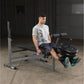 Man wearing gray t-shirt and dark shorts using gray and black weight bench with leg extension attachment and barbell plates, 