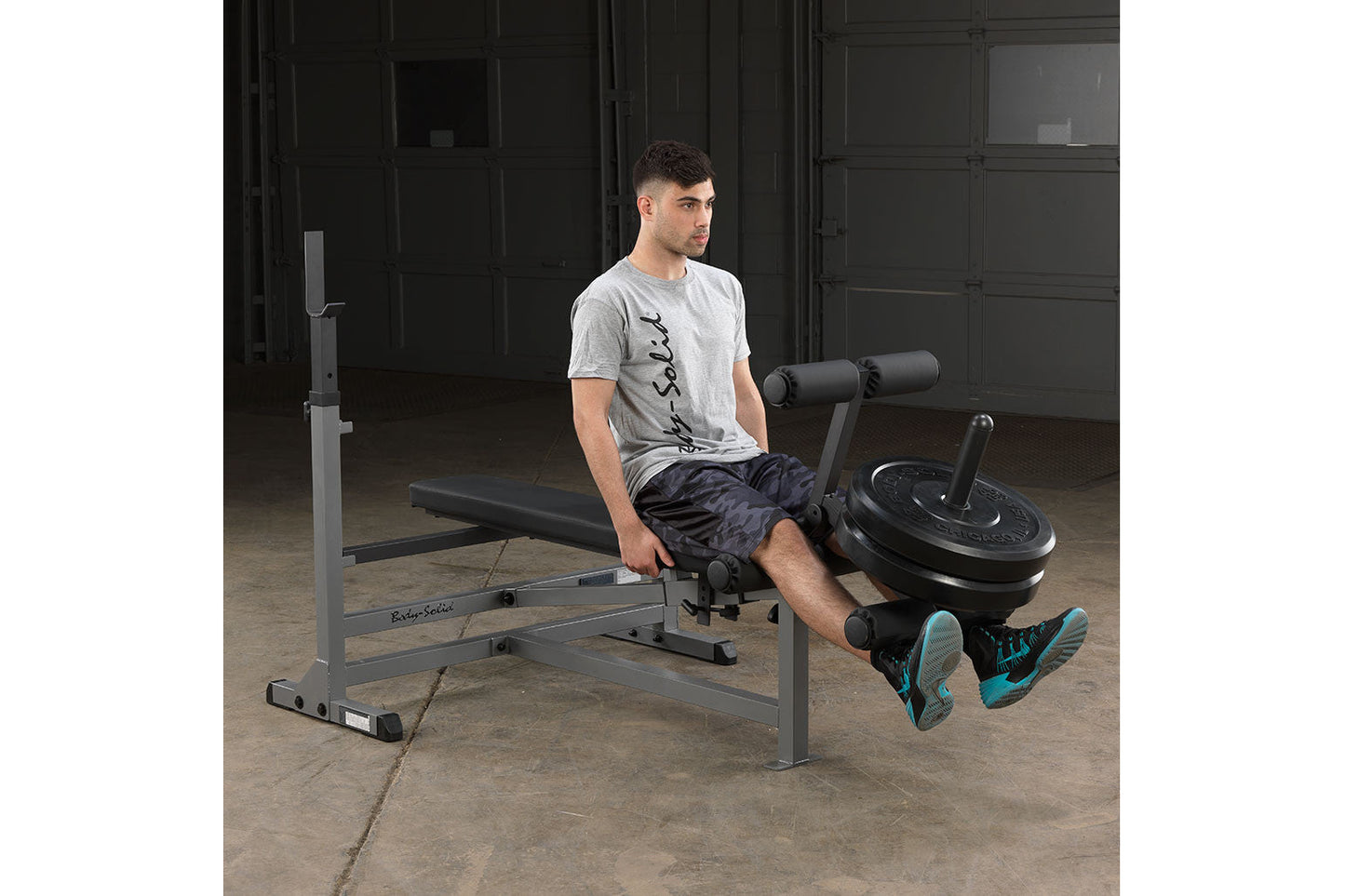 Man wearing gray t-shirt and dark shorts using gray and black weight bench with leg extension attachment and barbell plates, 