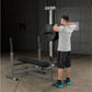 Man performing shoulder press with loaded barbell on gray and black weight bench with integrated rack in garage gym setting