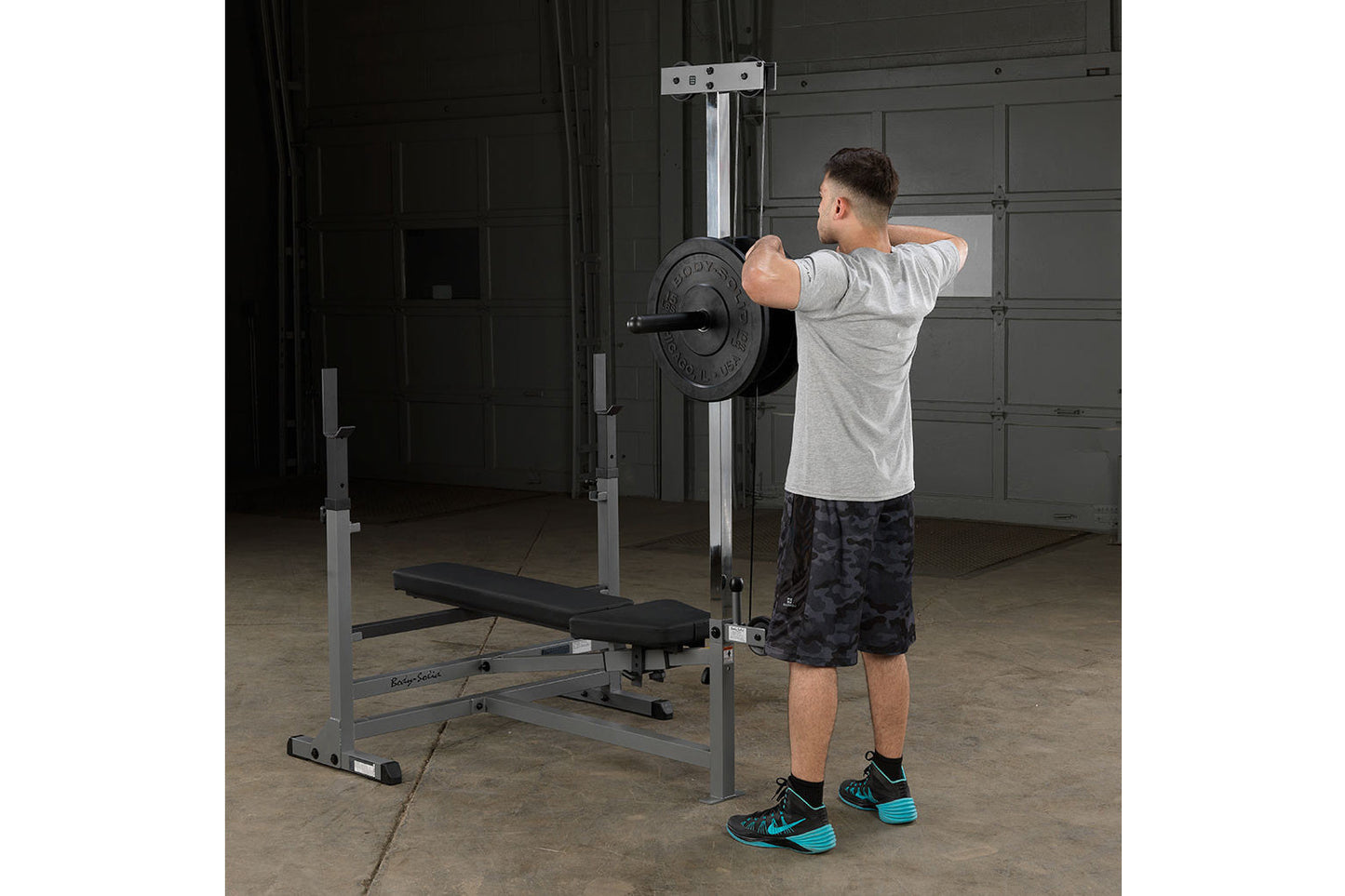 Man performing shoulder press with loaded barbell on gray and black weight bench with integrated rack in garage gym setting
