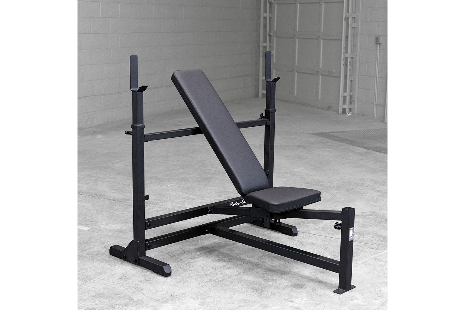 Black weight bench with incline backrest, leg extension attachment, and adjustable barbell uprights on concrete floor