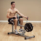 Shirtless man seated on gray and black leg press machine with weighted plate, demonstrating exercise form on wooden floor