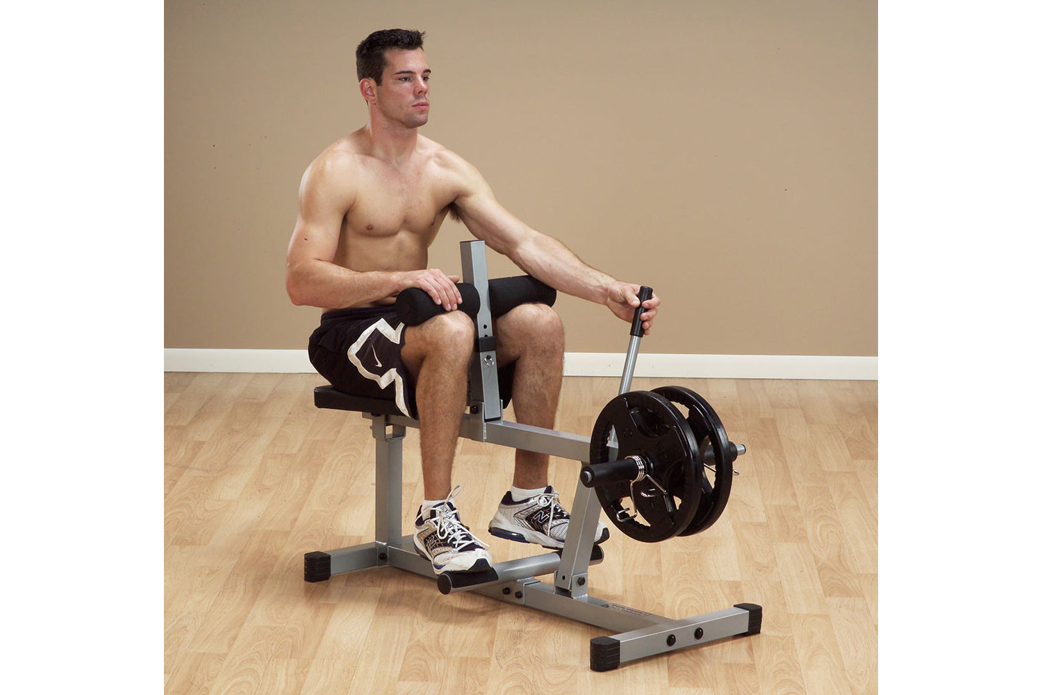 Shirtless man seated on gray and black leg press machine with weighted plate, demonstrating exercise form on wooden floor