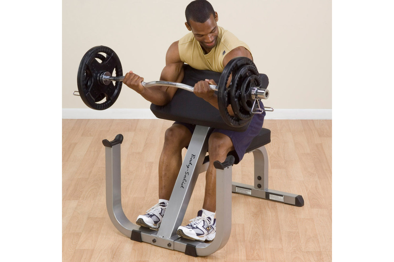 Man performing bicep curl on gray and black preacher curl bench with black weight plates and barbell, wearing yellow tank top