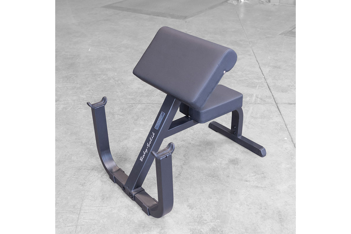 Body-Solid preacher curl bench with gray padded seat and backrest, black steel frame with leg extension, on concrete floor