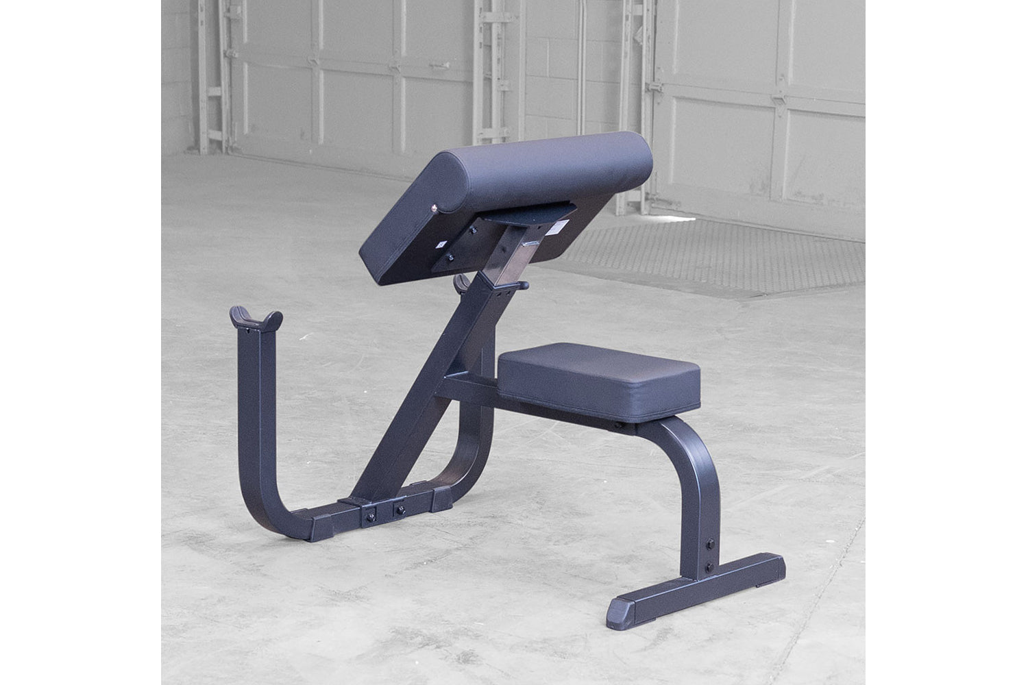 Navy blue preacher curl bench with padded seat and back support on concrete floor in gym setting