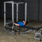 Man in blue shirt performing bench press exercise on white metal power rack with black barbell in industrial gym setting