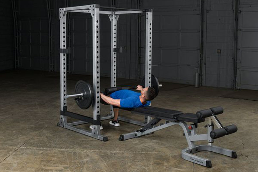 Man in blue shirt performing bench press exercise on white metal power rack with black barbell in industrial gym setting