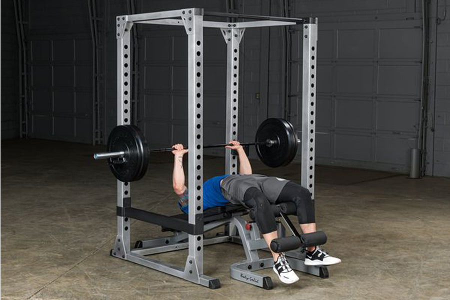 White metal power rack with adjustable safety bars and j-hooks, person performing barbell bench press in garage setting