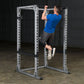 Man in blue shirt performing pull-up on silver power rack with black safety bars in gym setting