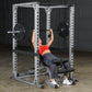 Person performing barbell bench press in white and black steel power rack with adjustable safety bars in gym setting