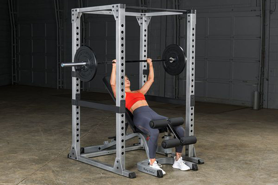 Person performing barbell bench press in white and black steel power rack with adjustable safety bars in gym setting