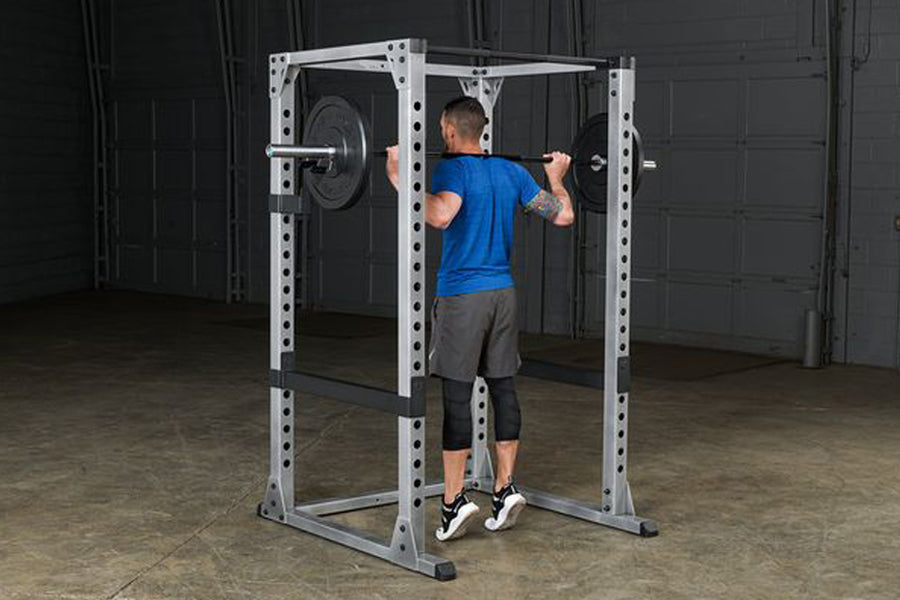 White and black steel power rack with man in blue shirt performing squat exercise, adjustable safety bars and weight loaded b