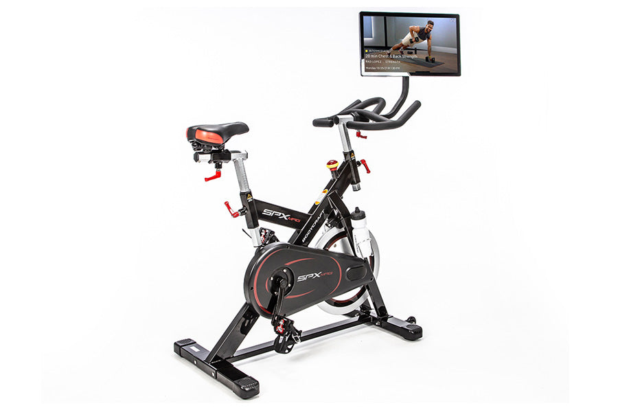 Black and red stationary indoor cycling bike with tablet mount displaying workout video, red handlebars, and padded seat on w