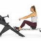 Woman using black BodyCraft rowing machine, wearing gray tank top and burgundy leggings, demonstrating proper form on white b