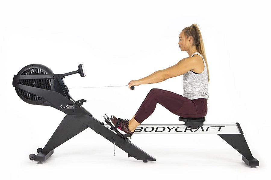 Woman using black BodyCraft rowing machine, wearing gray tank top and burgundy leggings, demonstrating proper form on white b