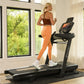 Woman in brown sports bra and orange leggings running on black BowFlex treadmill in bright home gym with windows