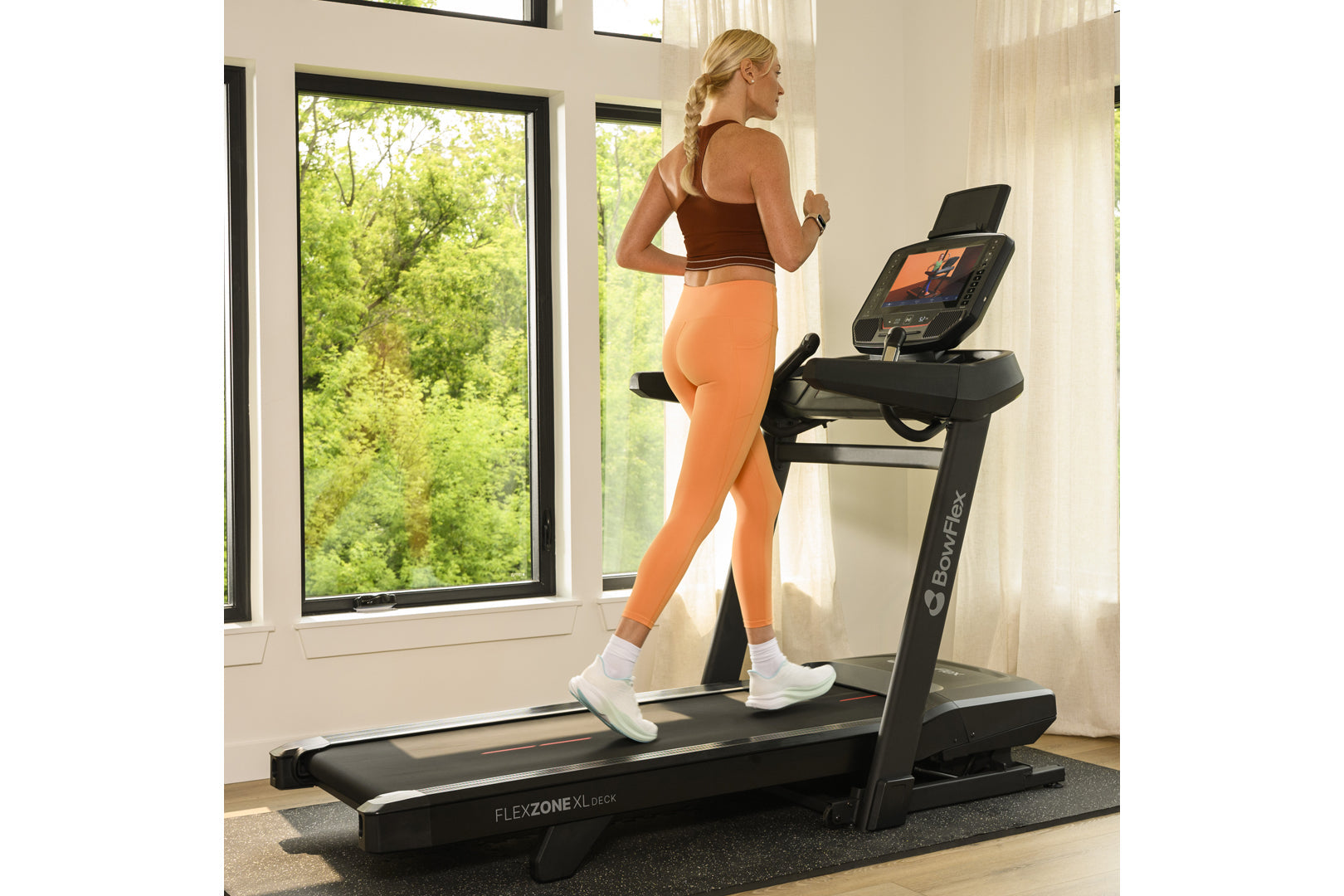 Woman in brown sports bra and orange leggings running on black BowFlex treadmill in bright home gym with windows