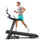 Man running on black Bowflex T6 treadmill, wearing gray sleeveless shirt and turquoise shorts with white sneakers, on white b