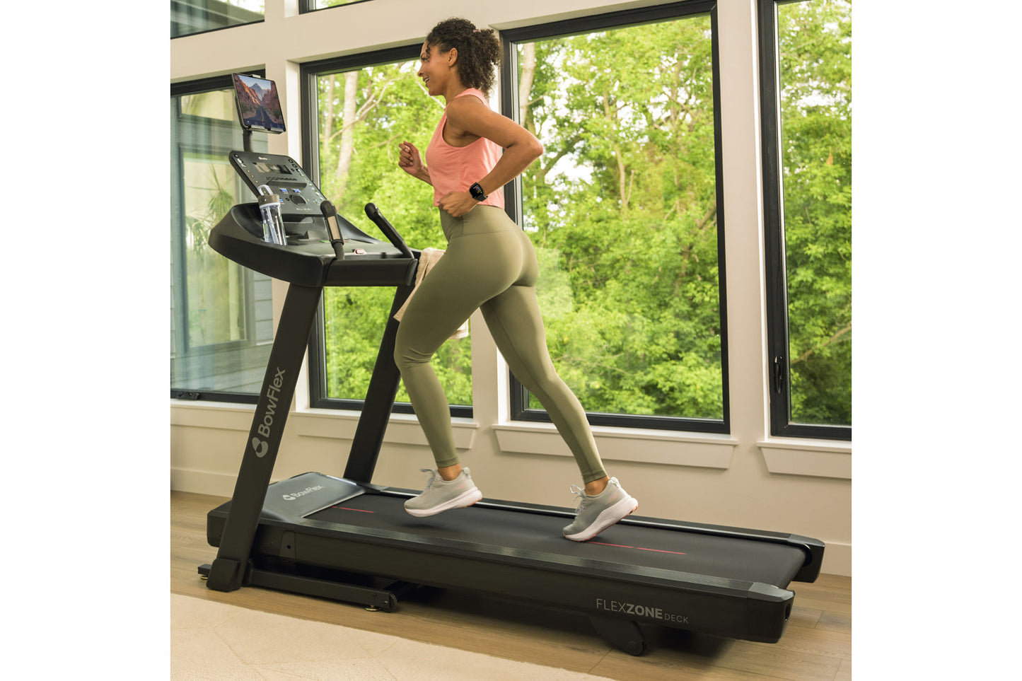 Woman wearing coral tank top and sage green leggings running on black Bowflex treadmill in bright gym with large windows