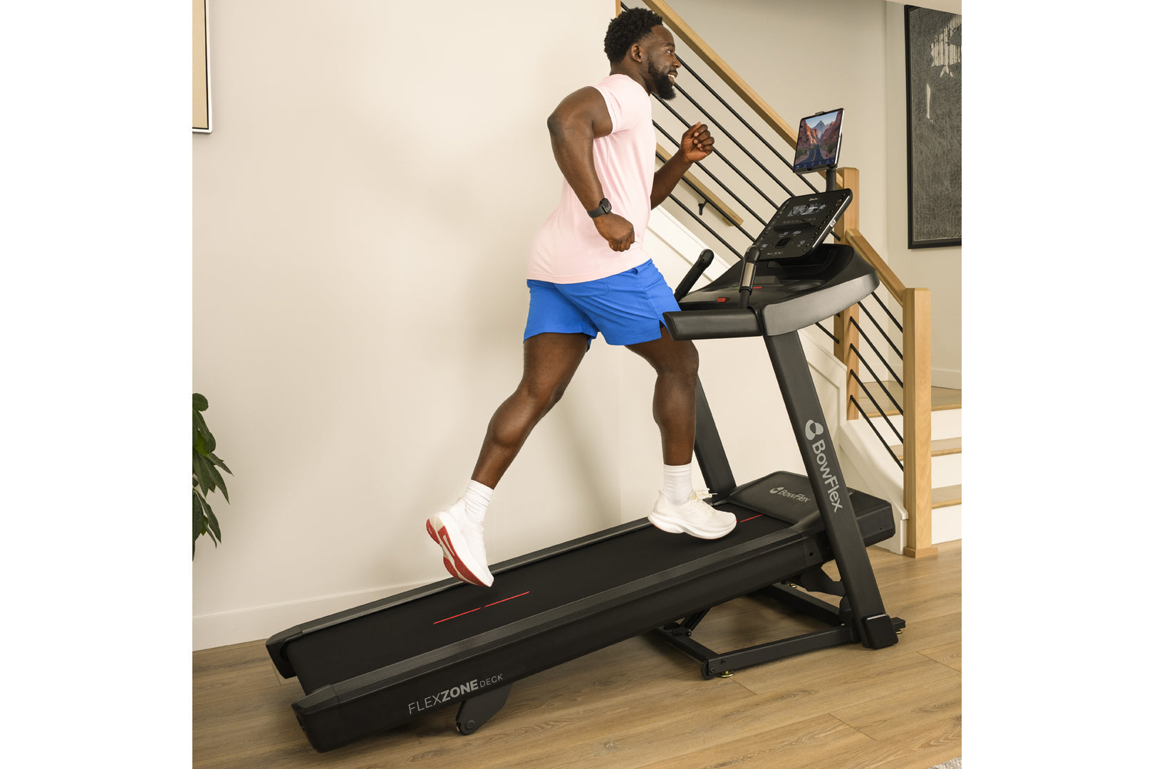 Man wearing light pink sleeveless tank top and blue shorts running on black Bowflex treadmill in home gym setting