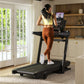 Woman running on black Bowflex treadmill wearing yellow sports bra and rust-orange leggings in modern home gym with wooden sh