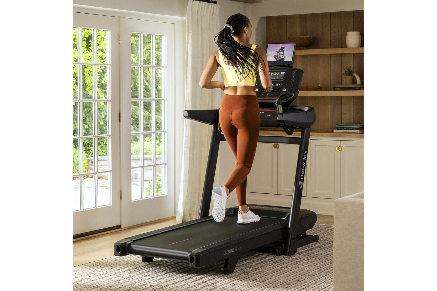 Woman running on black Bowflex treadmill wearing yellow sports bra and rust-orange leggings in modern home gym with wooden sh