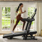 Woman running on Bowflex T9 treadmill wearing yellow sports bra and brown leggings in bright home gym with windows